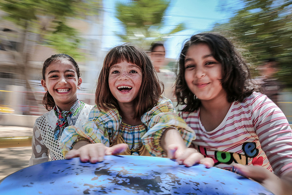 Three smiling Palestinian girls standing together outdoors, representing the human dimension of the Israeli-Palestinian conflict.